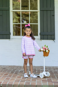 A young girl stands on a brick porch in front of a white wall with dark green shutters, wearing a light pink and white patterned long-sleeve shirt and matching shorts, holding a small basket of flowers next to a white scooter.