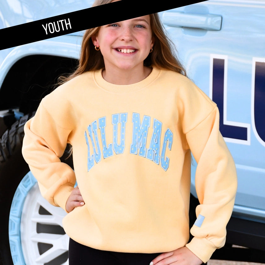 Young girl wearing a yellow 'LULU MAC' sweatshirt in front of a light blue vehicle.