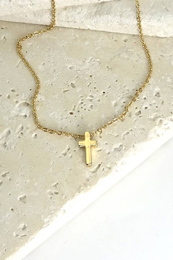 A delicate gold chain necklace with a small cross pendant, displayed on a textured light beige stone surface.