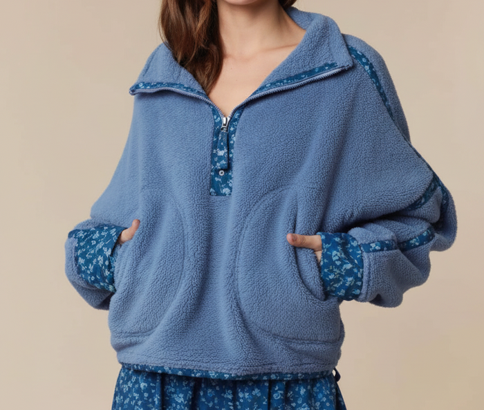 A woman modeling a dusty blue fleece half-zip pullover with blue floral patterned trim and matching floral skirt against a solid beige background.