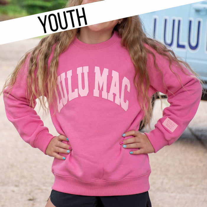 A young girl wearing a bright pink crewneck sweatshirt with 'LULU MAC' printed in white arched letters across the chest. The image is styled as a lifestyle shot with a dirt ground background and a white vehicle, featuring a 'YOUTH' banner overlay in the top left corner.