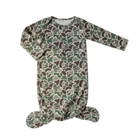 A baby footed sleeper with long sleeves, laid flat on a white wooden plank background. The sleeper features an all-over camouflage pattern in olive green, brown, and beige.