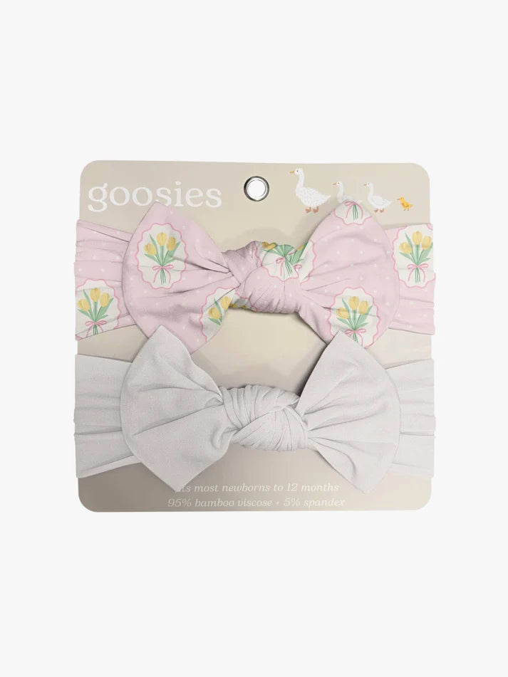 A two-piece baby headband set by Goosies, featuring a light pink headband with a yellow tulip pattern and a large bow, paired with a white headband and a large white bow, displayed on its cardboard backing.