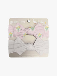A two-piece baby headband set by Goosies, featuring a light pink headband with a yellow tulip pattern and a large bow, paired with a white headband and a large white bow, displayed on its cardboard backing.