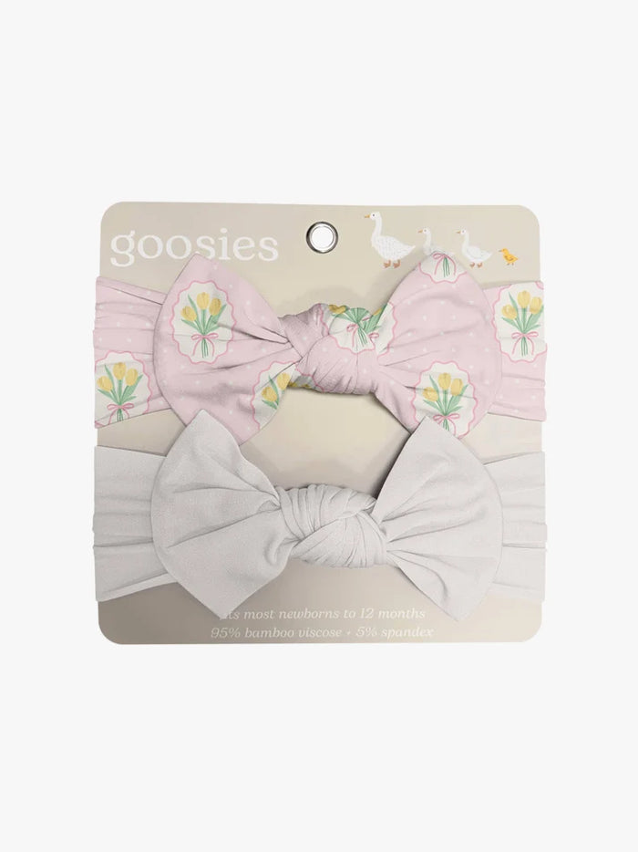 A two-piece baby headband set by Goosies, featuring a light pink headband with a yellow tulip pattern and a large bow, paired with a white headband and a large white bow, displayed on its cardboard backing.
