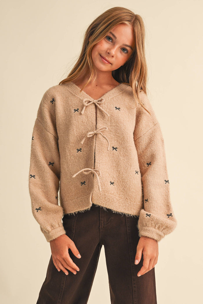 A young girl with long blonde hair wearing a fuzzy beige cropped cardigan with small black bow details and dark brown pants, posing against a solid light beige background.