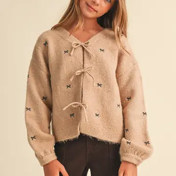 A young girl wearing a beige fuzzy knit cardigan sweater with bow details and drawstrings, paired with dark brown pants, against a solid cream background.