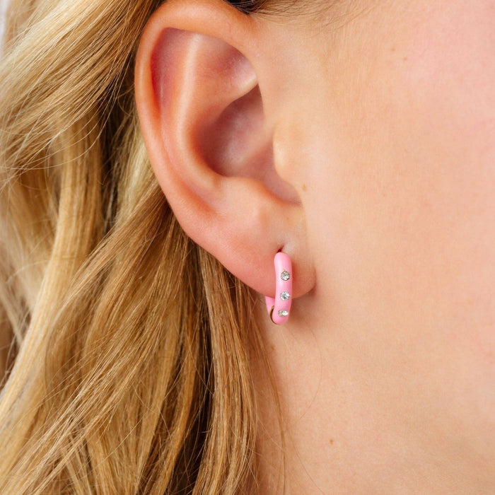 A close-up of a person's ear wearing a small pink huggie hoop earring decorated with tiny clear rhinestones.