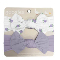 A two-piece baby headband set by Goosies, featuring a white headband with a purple and green berry print and a lavender-purple knotted bow headband, both on a beige backing card.