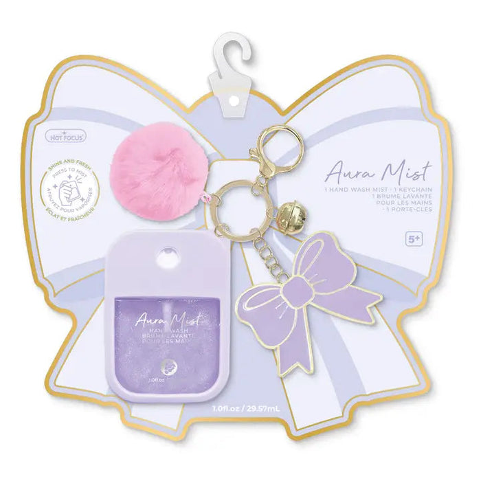 Hot Focus Aura Mist hand wash mist keychain set in a butterfly-shaped packaging card. The set includes a lavender hand wash mist bottle, a pink fluffy pom-pom, a gold-tone keychain ring, a small gold bell, and a lavender bow-shaped keychain.