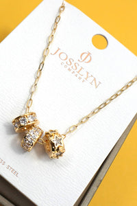 A gold-tone charm necklace with three charms, displayed on a white Josslyn Company card over a yellow background. The charms include two pavé-set clear stones and one star-shaped charm.