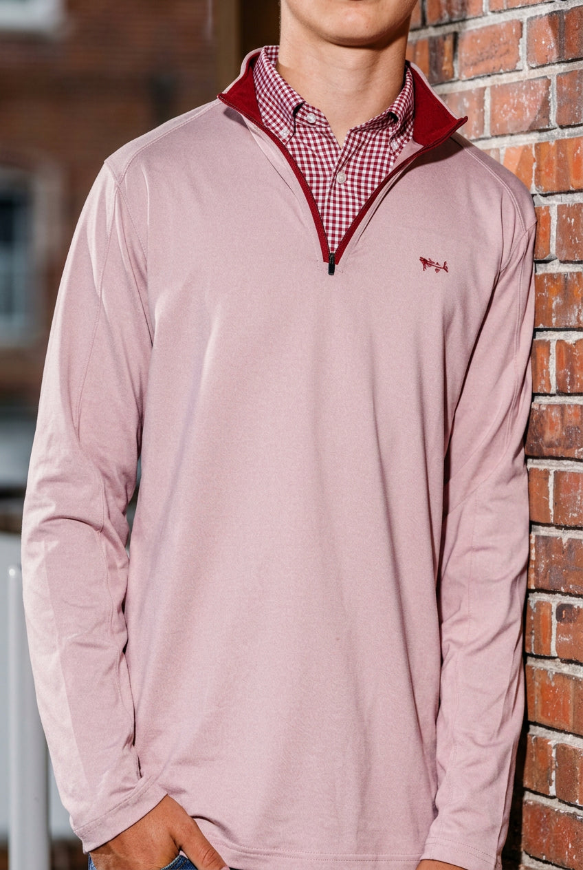 A man wearing a light pink quarter-zip pullover with a red inner collar, layered over a red and white checkered shirt, standing against a brick wall.