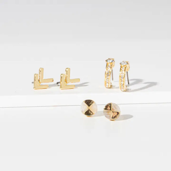 Four pairs of gold-toned stud earrings arranged on a white surface against a white background. The set includes two geometric L-shaped earrings, two curved pavé earrings with clear stones, and two round pavé earrings.