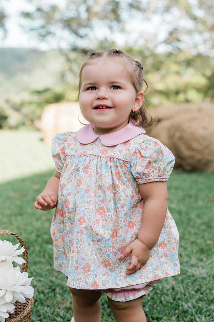 A toddler girl wearing a short-sleeve floral dress with a pink Peter Pan collar, standing outdoors on a grassy lawn with a blurred background of trees and hay bales.