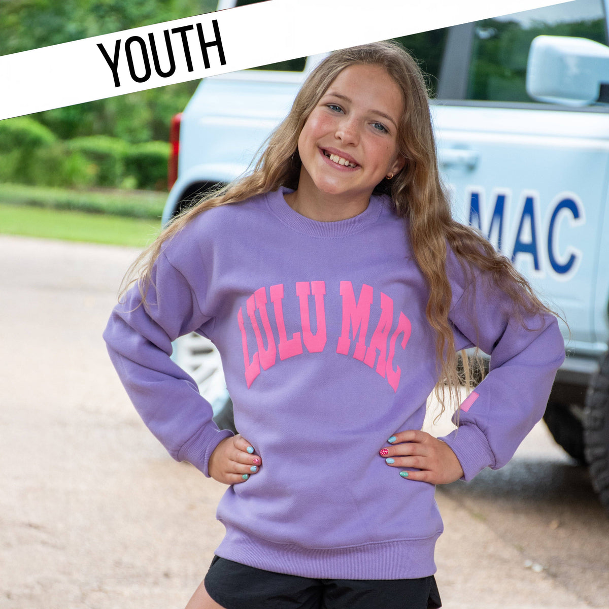 Young girl wearing a purple 'Lulu Mac' sweatshirt with a vehicle in the background