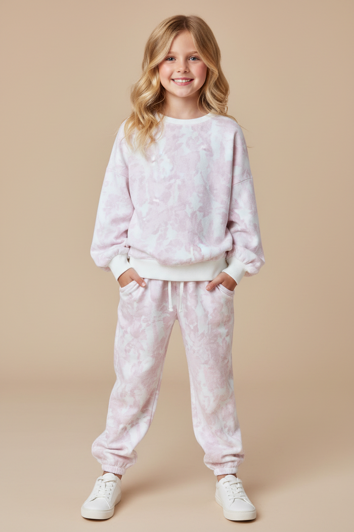 A young girl with blonde hair stands against a solid beige background wearing a matching light pink and white tie-dye sweatshirt and jogger pants set, paired with white sneakers.