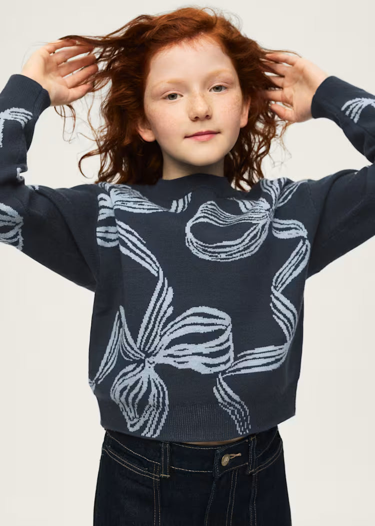 A young girl with curly red hair wearing a navy blue knit sweater with a white abstract bow pattern, paired with dark blue jeans, posing against a light gray background.