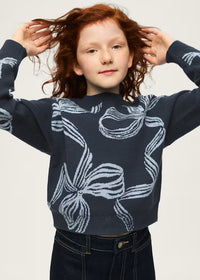 A young girl with curly red hair wearing a navy blue knit sweater with a white abstract bow pattern, paired with dark blue jeans, posing against a light gray background.