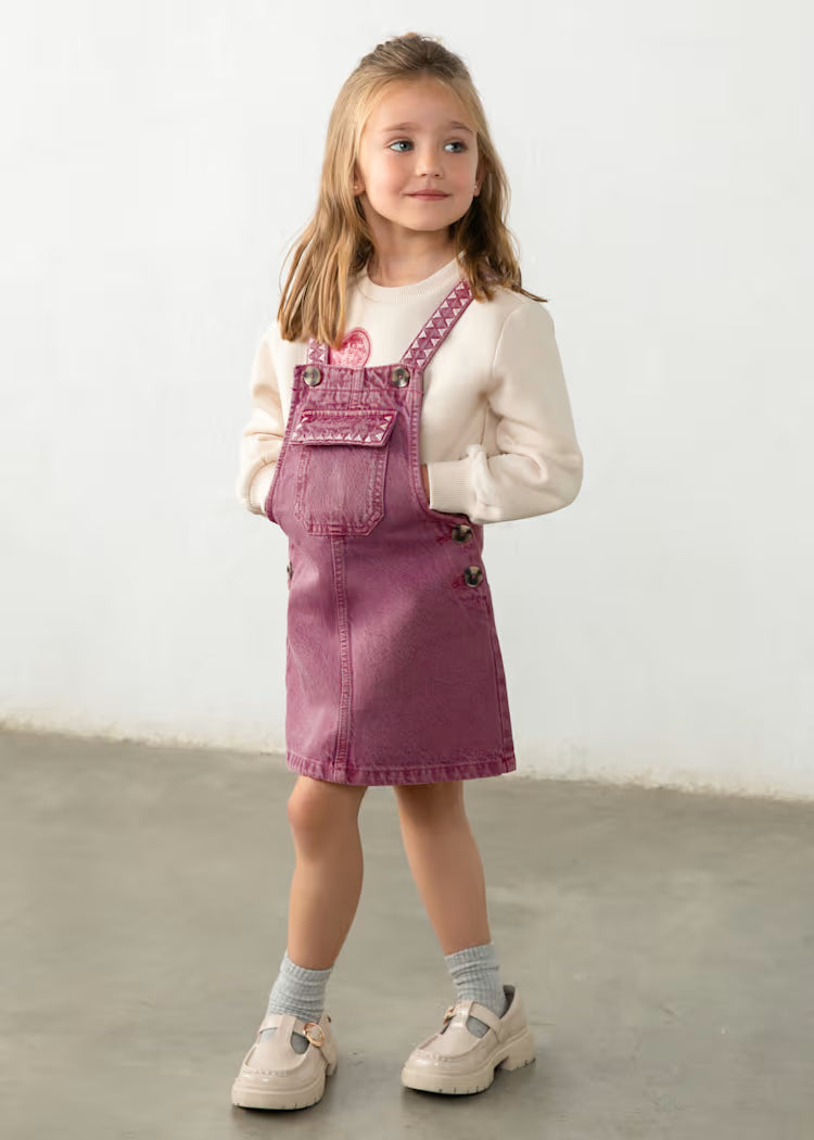 A young girl stands in a studio wearing a dusty rose denim overall mini dress over a cream long-sleeve shirt, paired with gray socks and cream Mary Jane shoes.