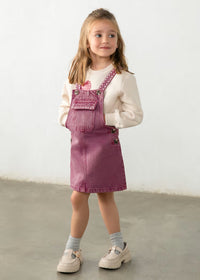 A young girl stands in a studio wearing a dusty rose denim overall mini dress over a cream long-sleeve shirt, paired with gray socks and cream Mary Jane shoes.
