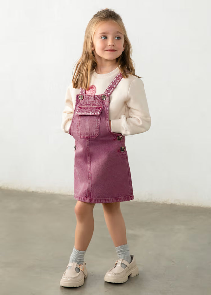 A young girl stands in a studio wearing a dusty rose denim overall mini dress over a cream long-sleeve shirt, paired with gray socks and cream Mary Jane shoes.