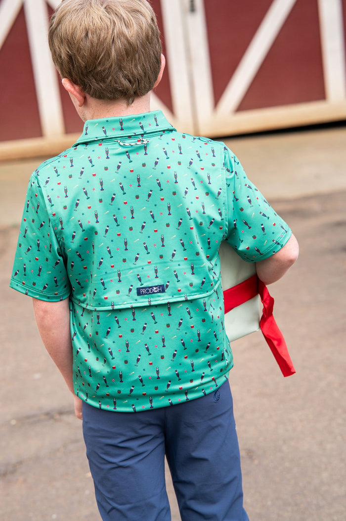 A child viewed from behind wearing a mint green short-sleeve collared shirt with a small repeating red and black pattern, paired with navy pants. The child is standing outdoors in front of a red barn door, holding a white tote bag with a red ribbon.
