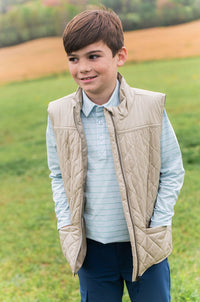 A young boy wearing a beige quilted zip-front vest over a light blue striped polo shirt and navy pants, standing in a grassy outdoor field.
