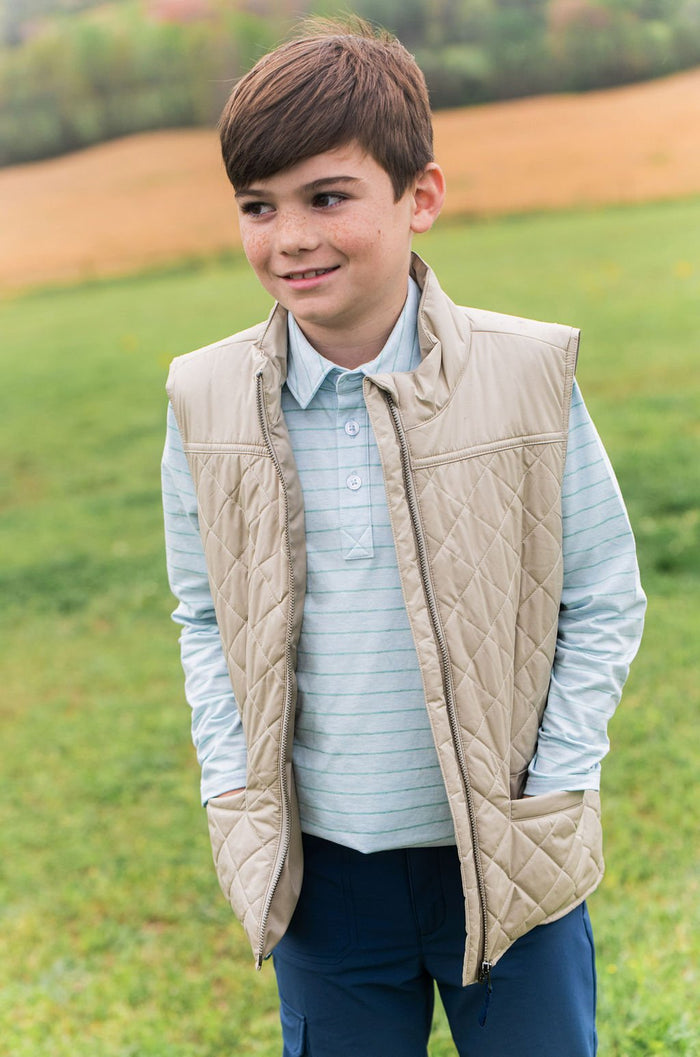 A young boy wearing a beige quilted zip-front vest over a light blue striped polo shirt and navy pants, standing in a grassy outdoor field.