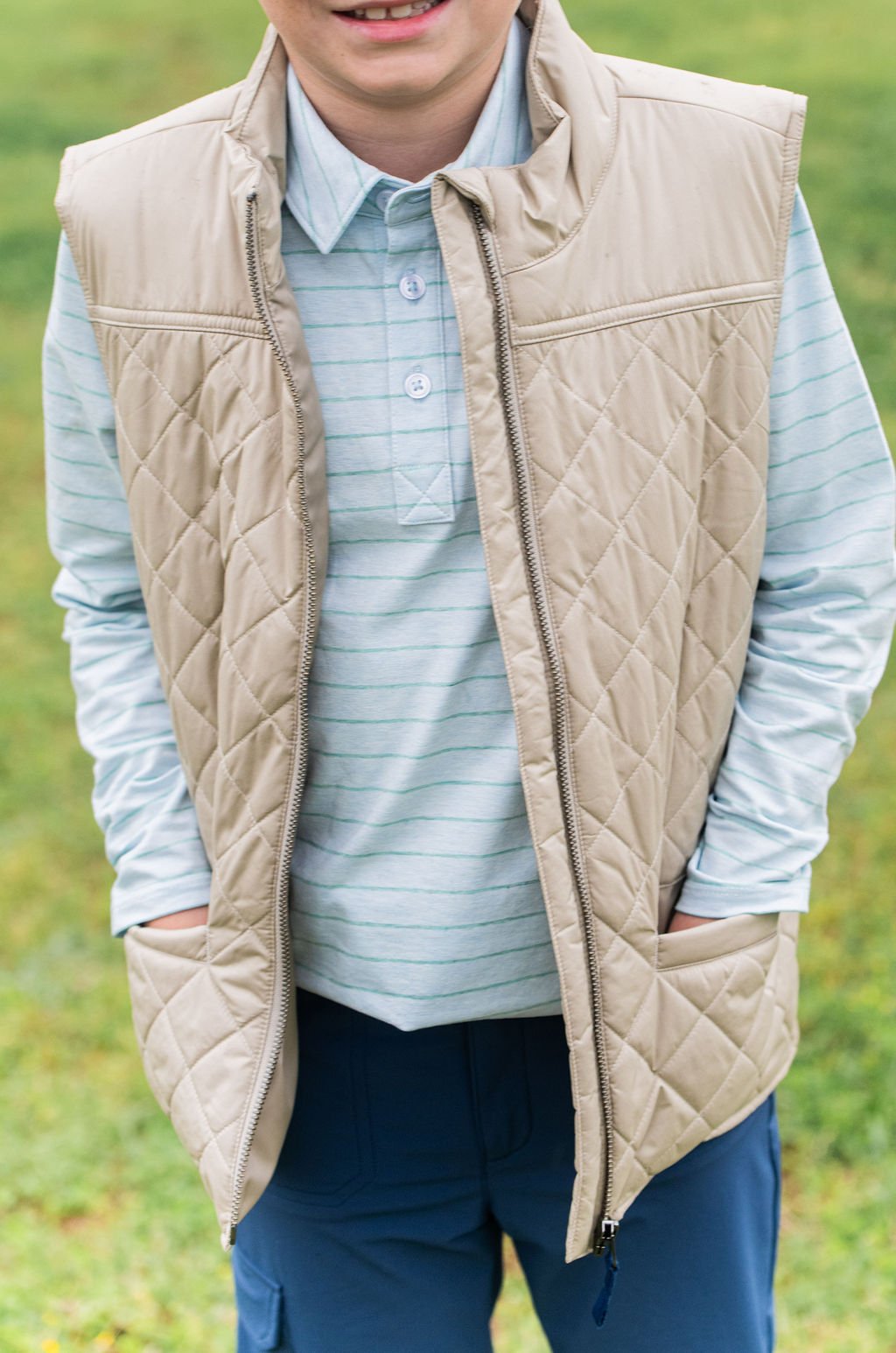 A person wearing a beige quilted zip-front vest over a light blue striped button-up shirt and navy pants, standing outdoors against a blurred grassy background.