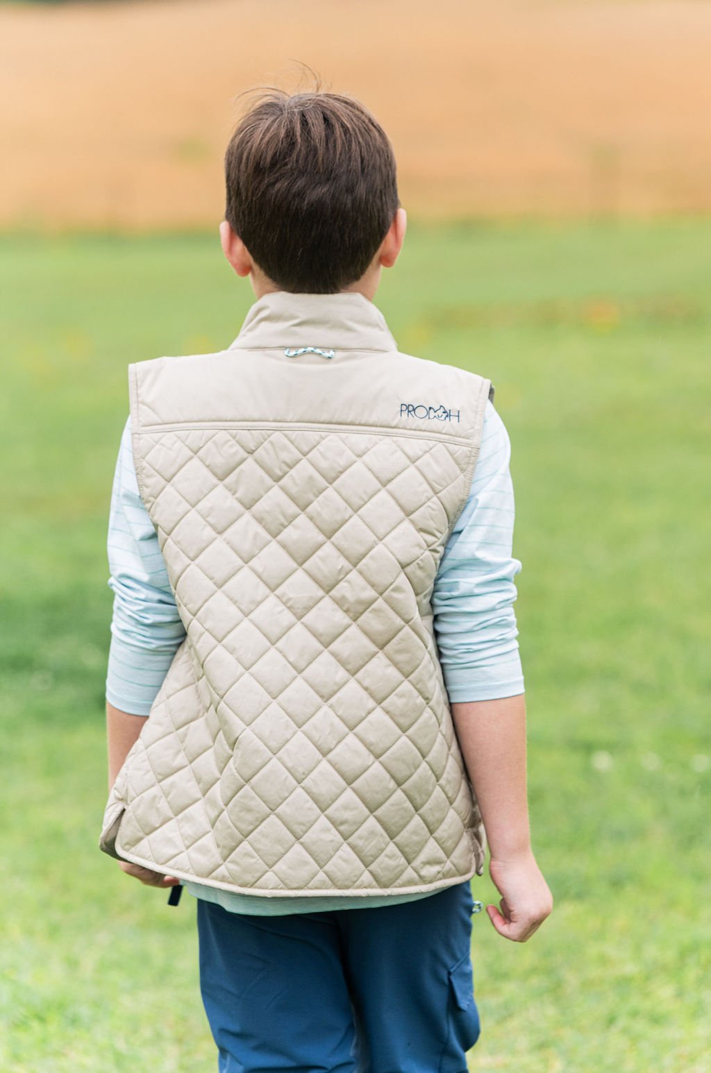 A child viewed from behind wearing a light beige diamond-quilted vest over a light blue long-sleeve shirt and blue pants, standing outdoors on a grassy field.