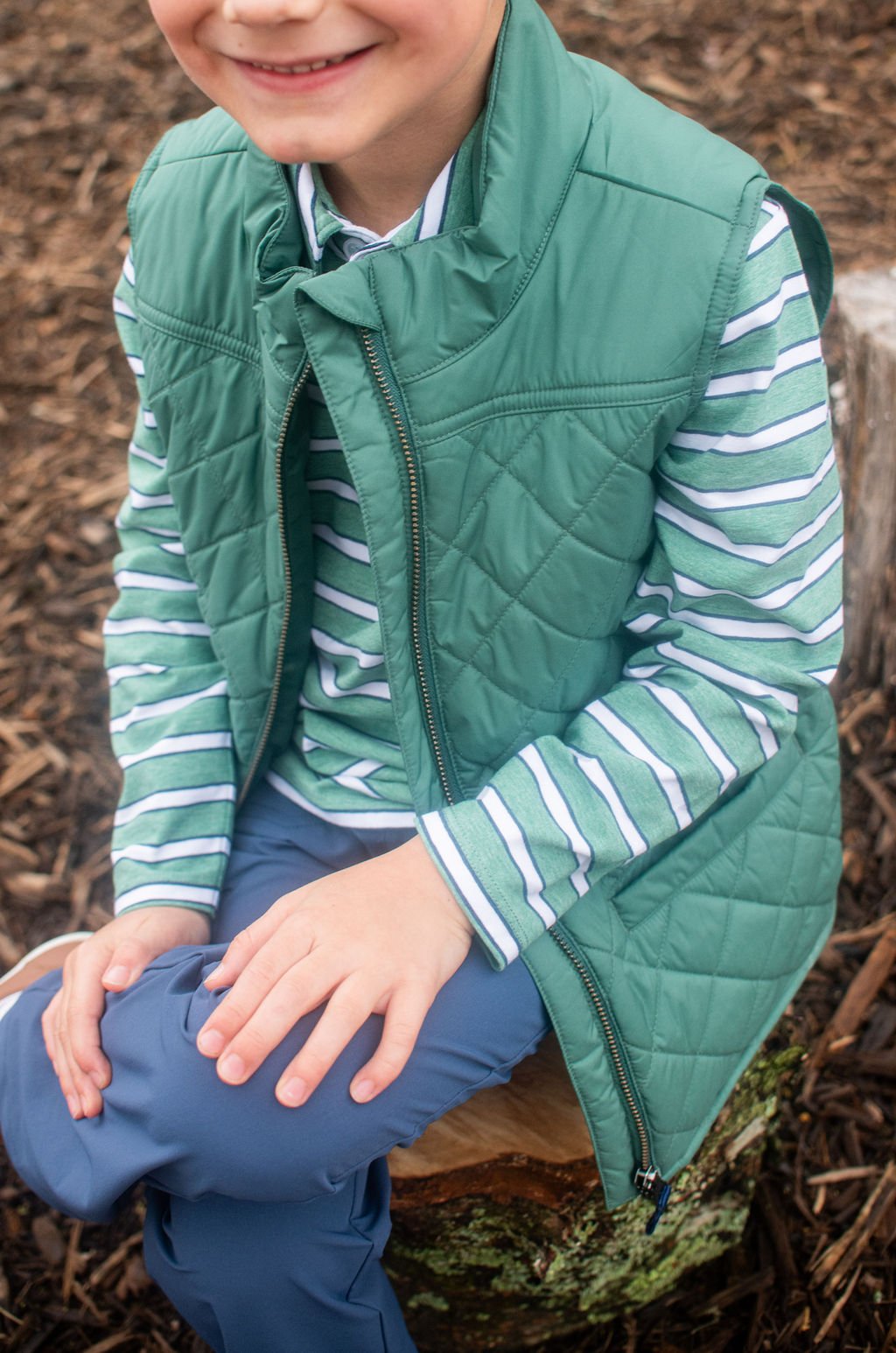 Child wearing a green quilted vest over a striped shirt sitting on a log outdoors.