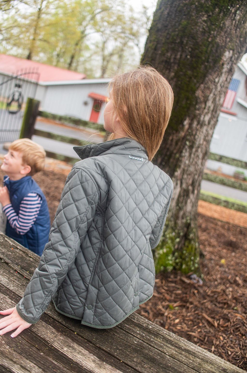 Child wearing a gray quilted jacket sitting on a wooden bench outdoors.