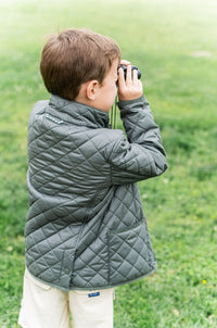 Child in a quilted jacket using binoculars in a grassy field