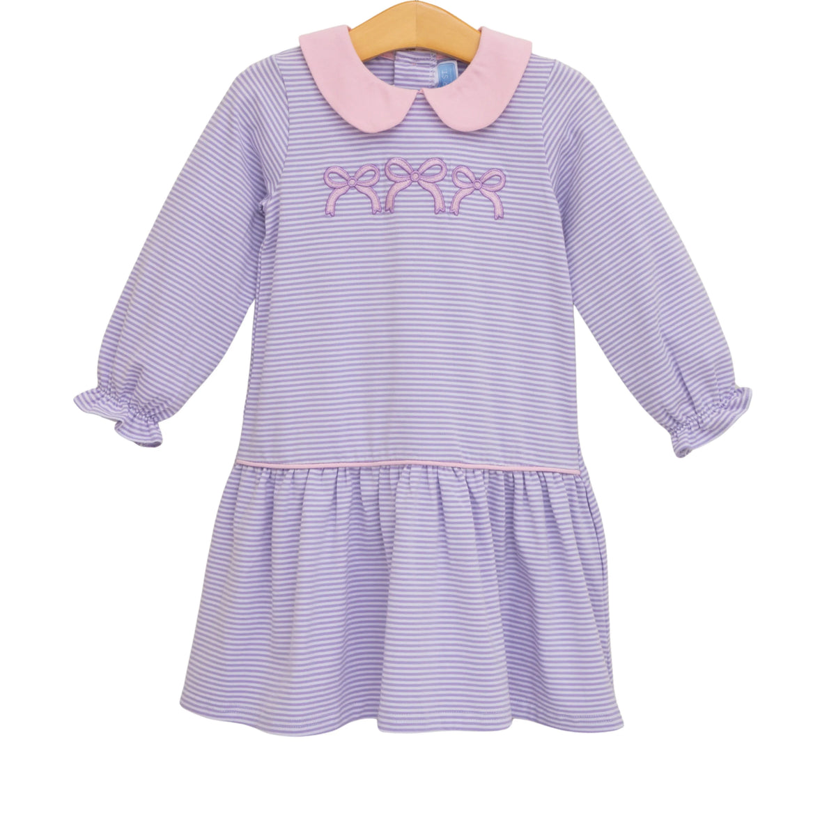 A children's long-sleeve dress with lavender and white horizontal stripes, a light pink Peter Pan collar, and pink embroidered bows across the chest. The dress has a ruffled hem and gathered cuffs, hanging on a wooden hanger against a plain white background.