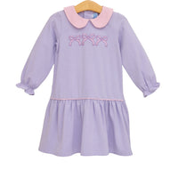 A children's long-sleeve dress with lavender and white horizontal stripes, a light pink Peter Pan collar, and pink embroidered bows across the chest. The dress has a ruffled hem and gathered cuffs, hanging on a wooden hanger against a plain white background.