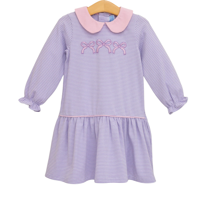 A children's long-sleeve dress with lavender and white horizontal stripes, a light pink Peter Pan collar, and pink embroidered bows across the chest. The dress has a ruffled hem and gathered cuffs, hanging on a wooden hanger against a plain white background.