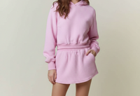 A woman modeling a light pink ribbed knit hoodie and matching mini skirt set, paired with white cowboy boots, against a solid light beige studio background.