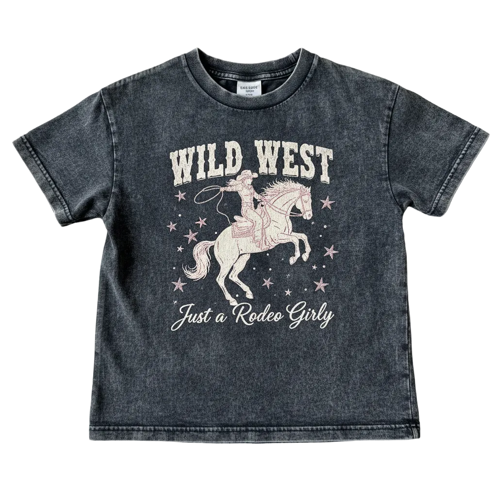 A kids' short-sleeve t-shirt in a washed charcoal gray color, featuring a pink and white graphic of a cowboy riding a horse with the text 'WILD WEST' and 'Just a Rodeo Girly'. The shirt is laid flat on a textured beige fabric background.