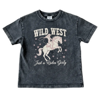 A kids' short-sleeve t-shirt in a washed charcoal gray color, featuring a pink and white graphic of a cowboy riding a horse with the text 'WILD WEST' and 'Just a Rodeo Girly'. The shirt is laid flat on a textured beige fabric background.