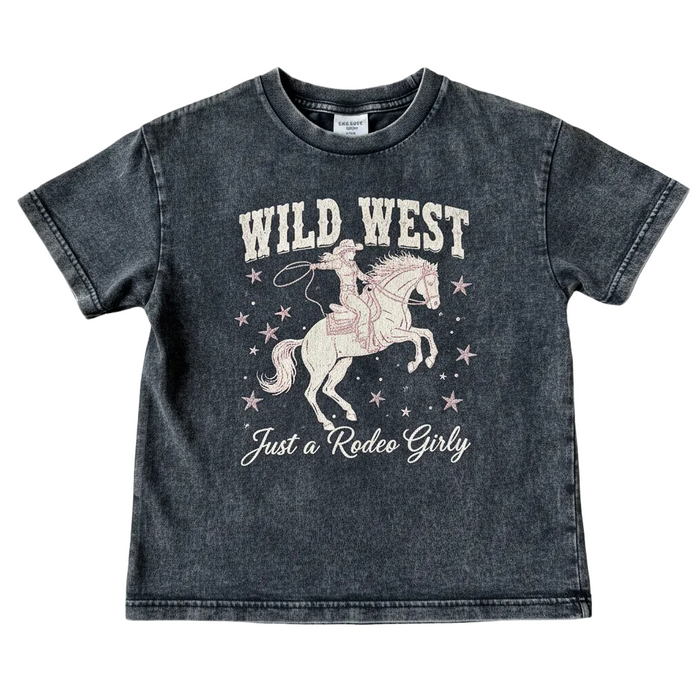 A kids' short-sleeve t-shirt in a washed charcoal gray color, featuring a pink and white graphic of a cowboy riding a horse with the text 'WILD WEST' and 'Just a Rodeo Girly'. The shirt is laid flat on a textured beige fabric background.
